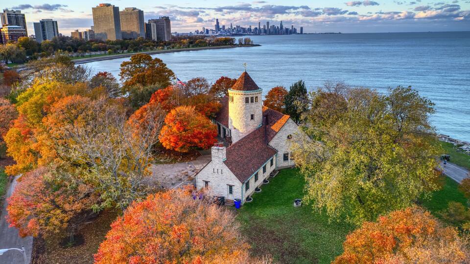 A historic building with a tower, surrounded by autumn foliage and a river.