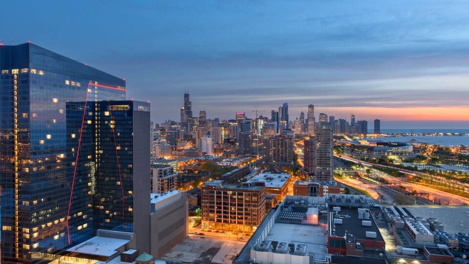 A cityscape at dusk with illuminated skyscrapers and a river in the background.