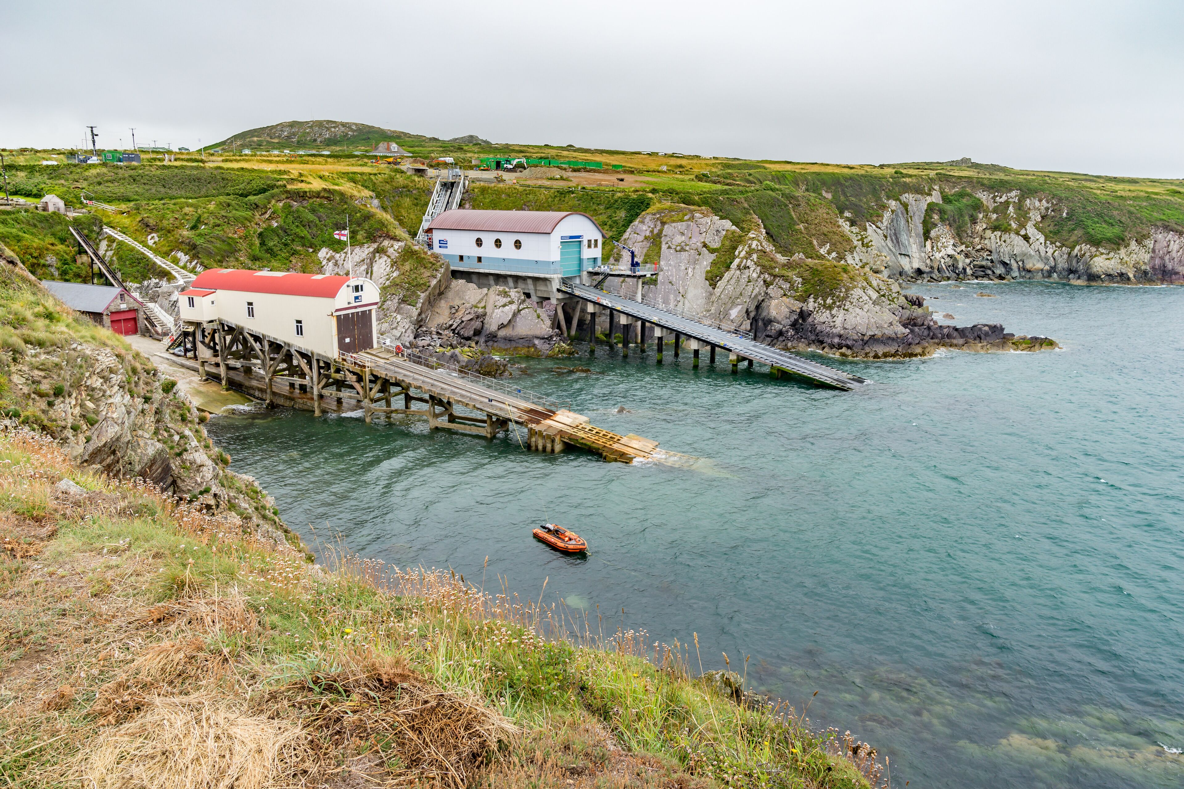 A coastal pier with a small boat, a white and blue building, and rocky cliffs.