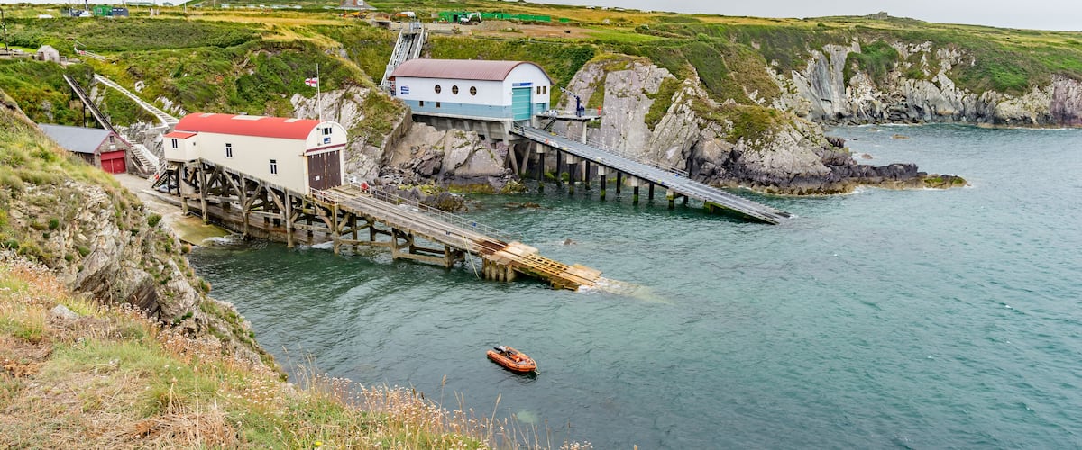 A coastal pier with a small boat, a white and blue building, and rocky cliffs.