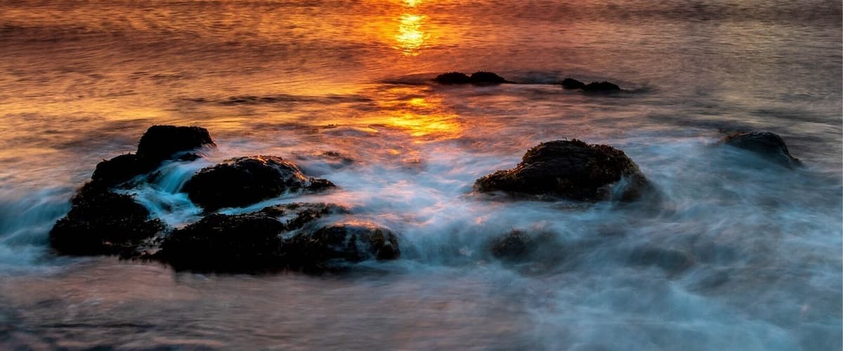 A sunset over a rocky shoreline with waves gently lapping at the rocks.