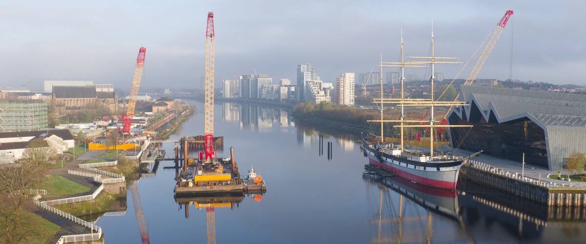 A river with a tall ship docked on the right and construction cranes on the left.