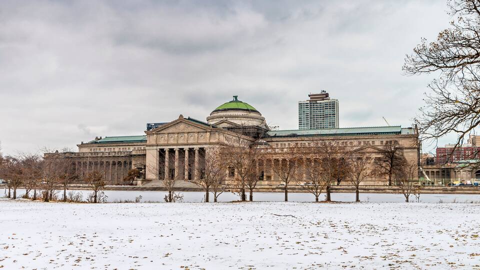 A large classical building with a green dome and columns, surrounded by snow-covered ground and leafless trees.