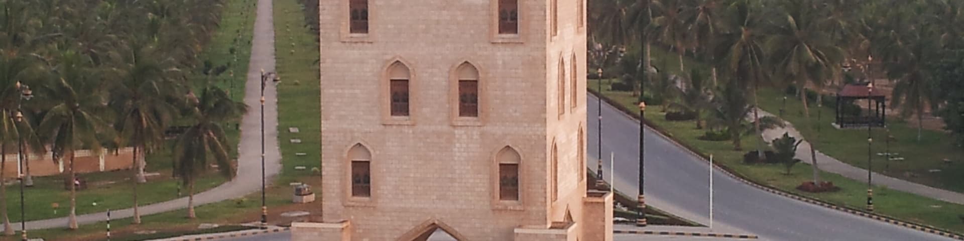 Um edifício grande e histórico com uma torre do relógio, rodeado por um jardim bem cuidado e palmeiras.