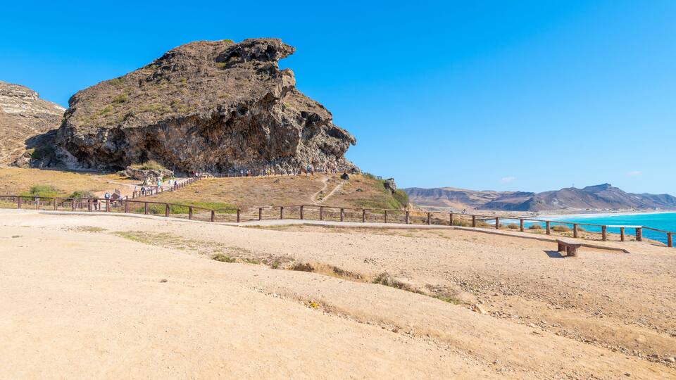 A rocky cliff with a wooden walkway leading up to it, overlooking a beach with clear blue water.