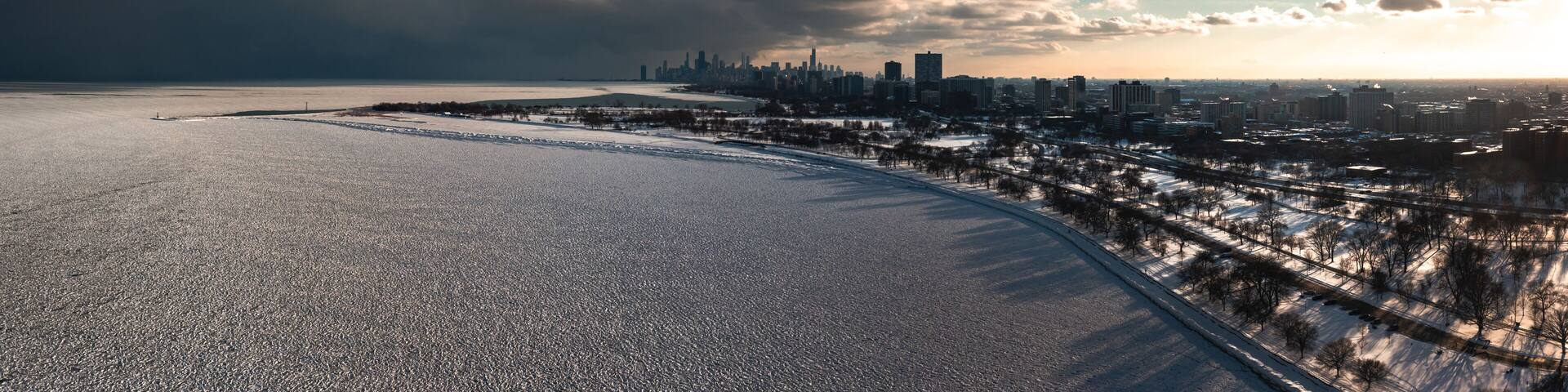 Aerial panorama of the Lake Michigan shoreline on the north side of Chicago covered in frozen ice 'pancakes' as clouds move past the downtown skyline in the distance and the sun begins to set.