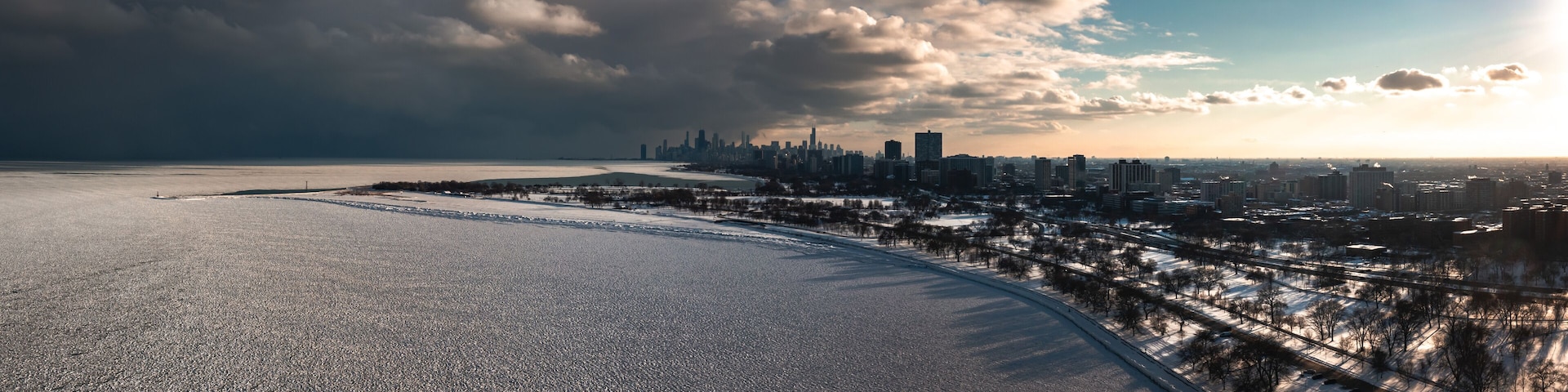 Aerial panorama of the Lake Michigan shoreline on the north side of Chicago covered in frozen ice 'pancakes' as clouds move past the downtown skyline in the distance and the sun begins to set.