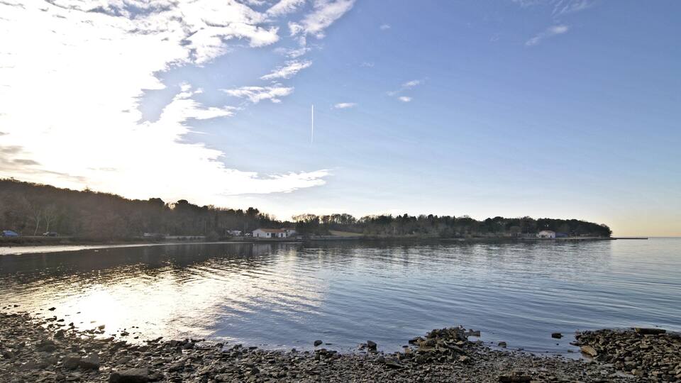 A calm lake with a rocky shoreline, a distant tree line, and a clear sky.