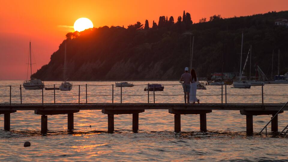 A sunset over a harbor with boats, a pier, and a hill in the background.