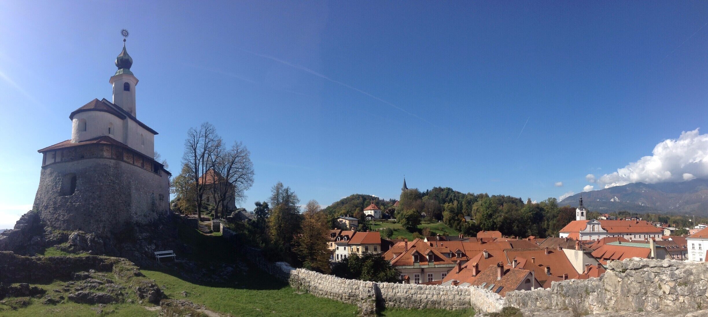A historic tower with a weather vane, surrounded by stone walls, and a village with red-roofed buildings in the background.