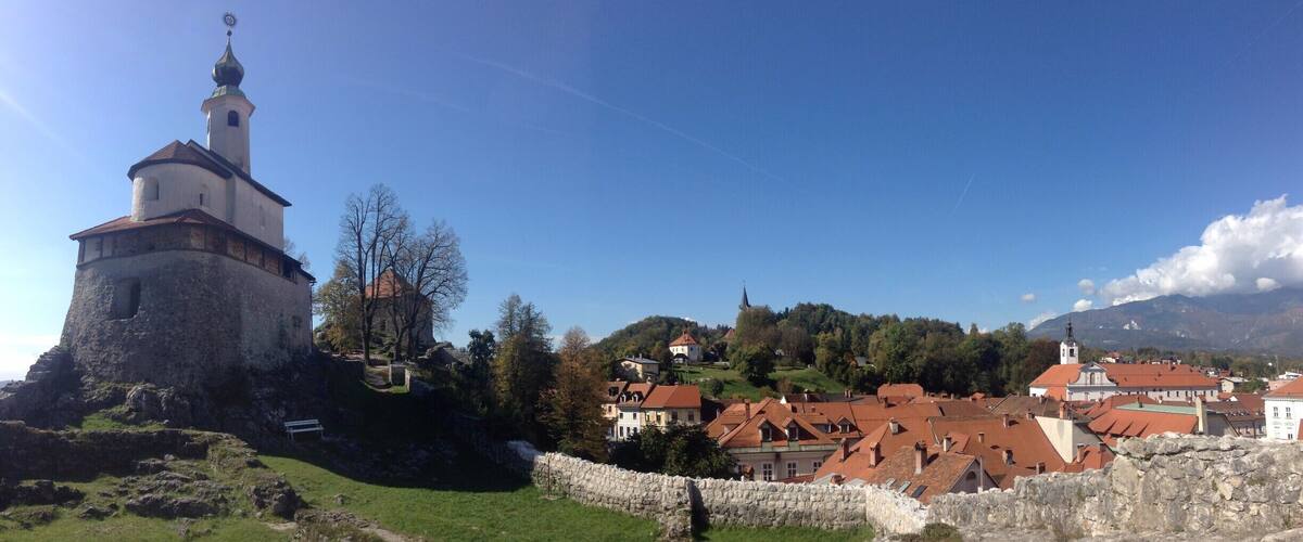 A historic tower with a weather vane, surrounded by stone walls, and a village with red-roofed buildings in the background.