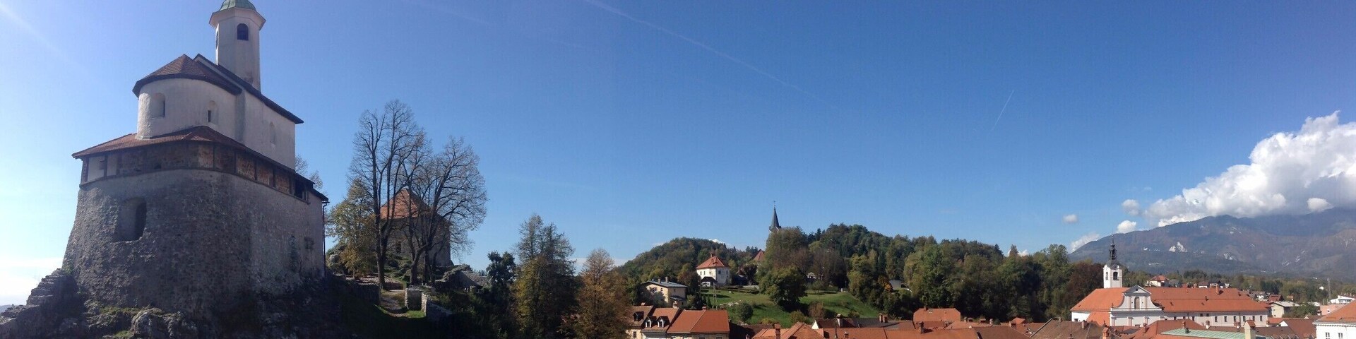A historic tower with a weather vane, surrounded by stone walls, and a village with red-roofed buildings in the background.