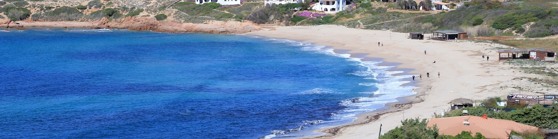 Paisagem costeira com praia arenosa, mar azul cristalino e casas espalhadas numa encosta.