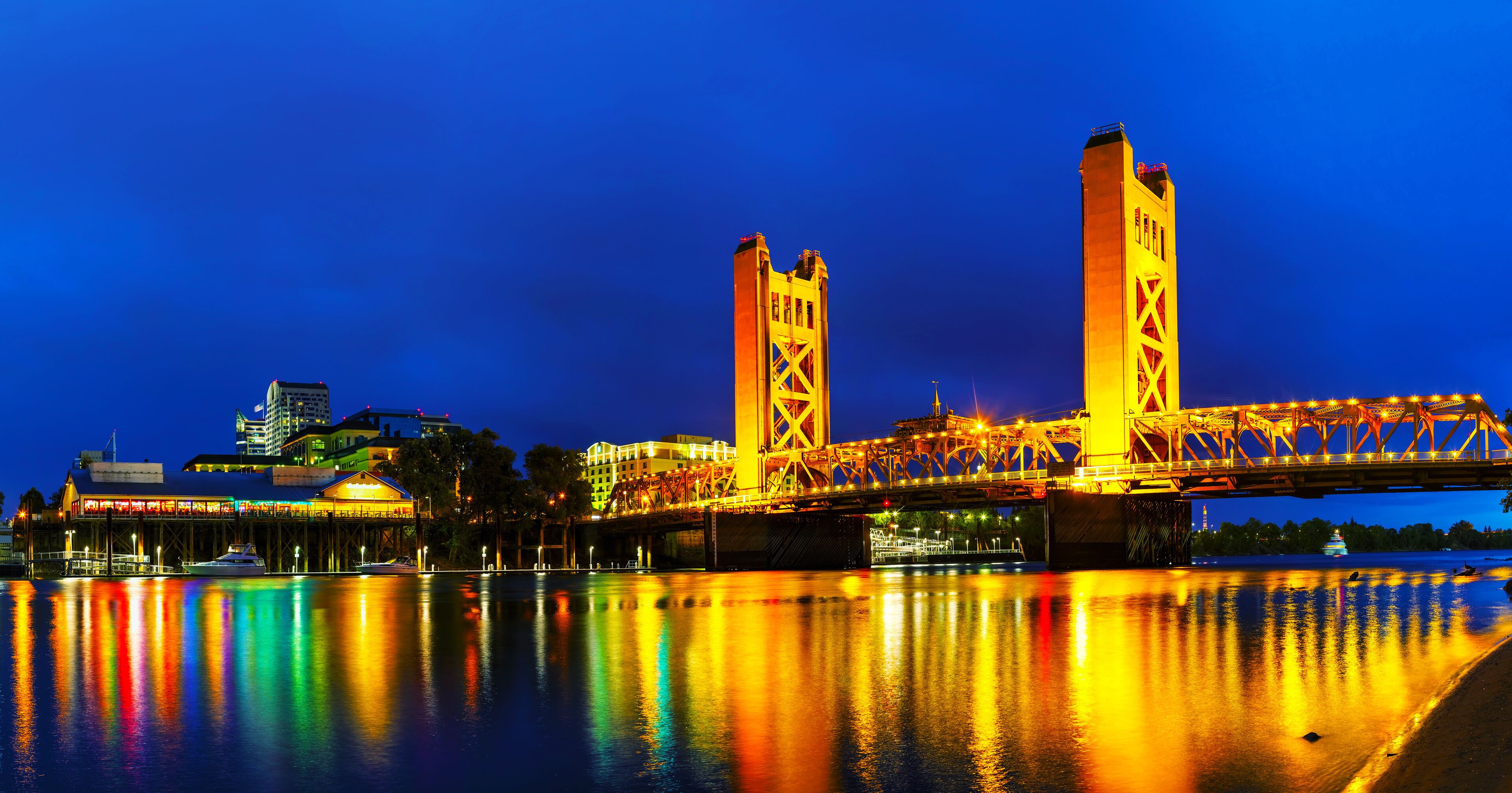 A brightly lit bridge with two towers reflecting on the water at night.