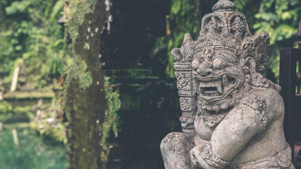 A stone statue of a mythical creature with intricate carvings, set against a backdrop of lush greenery and a moss-covered tree trunk.