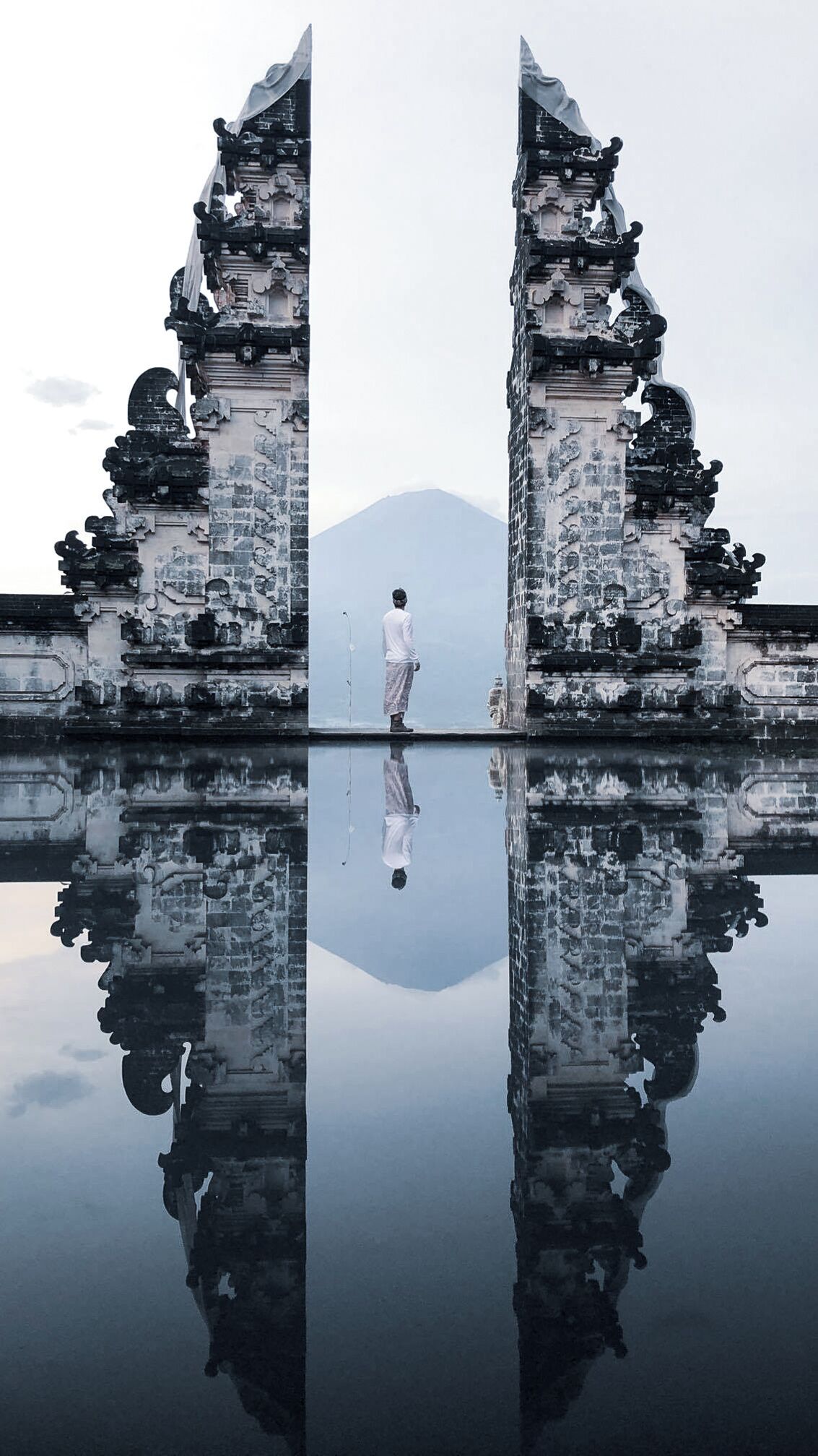 A man standing between two ancient, ornate gates reflecting in a calm body of water with a mountain in the background.