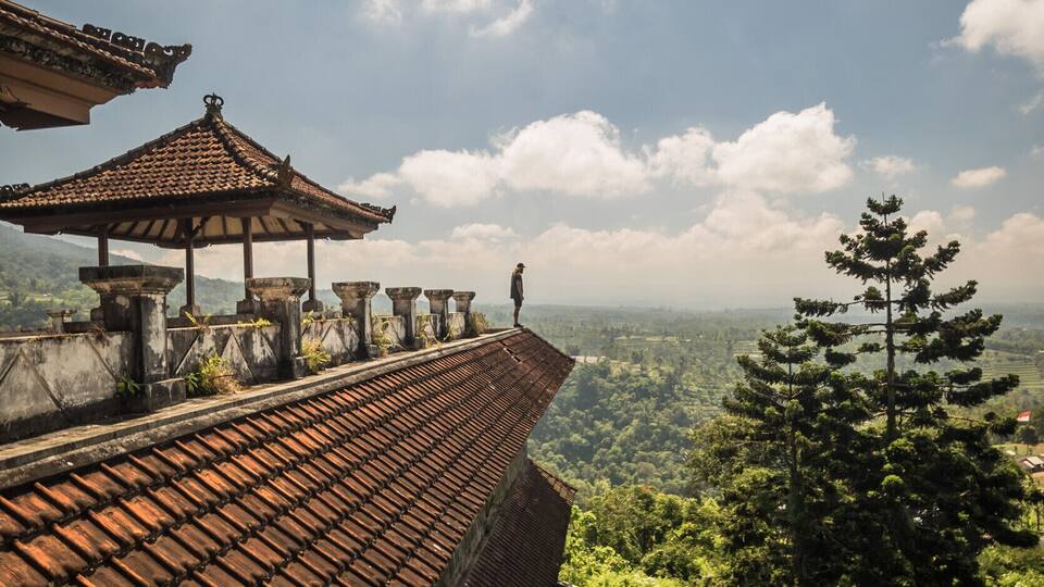 A traditional tiled roofed structure with a person standing on a ledge overlooking a lush green landscape.