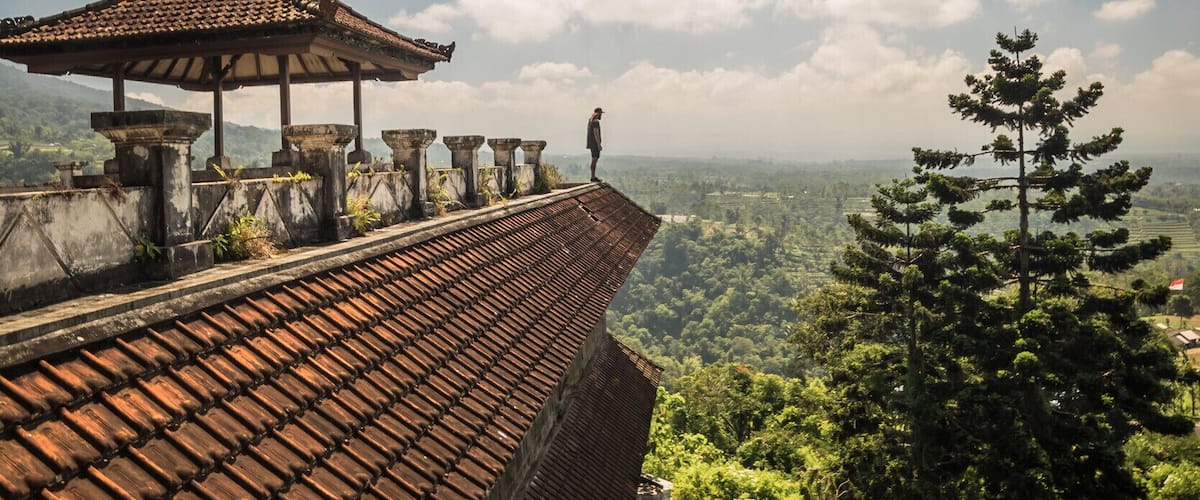 A traditional tiled roofed structure with a person standing on a ledge overlooking a lush green landscape.