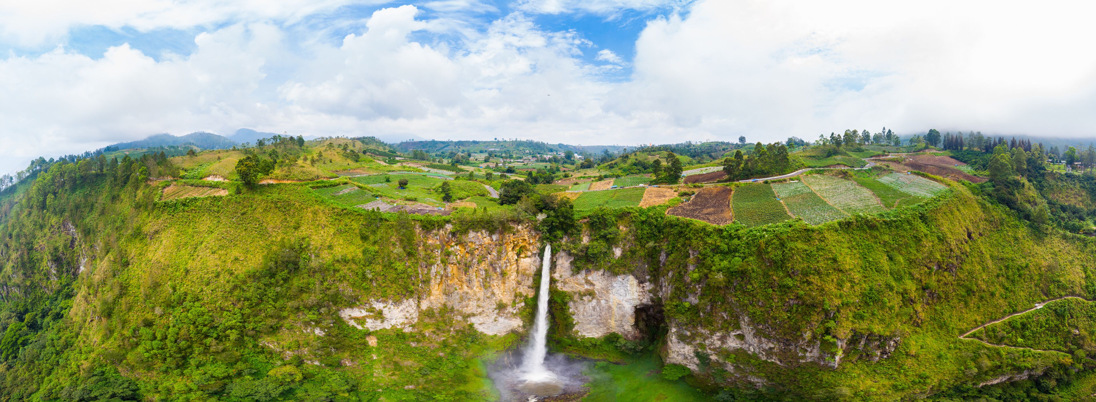 A lush green landscape with terraced fields, a waterfall, and a mountain range in the background.