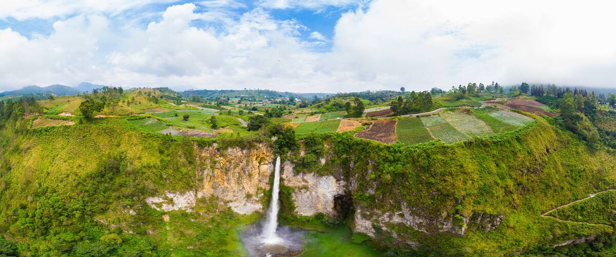 A lush green landscape with terraced fields, a waterfall, and a mountain range in the background.