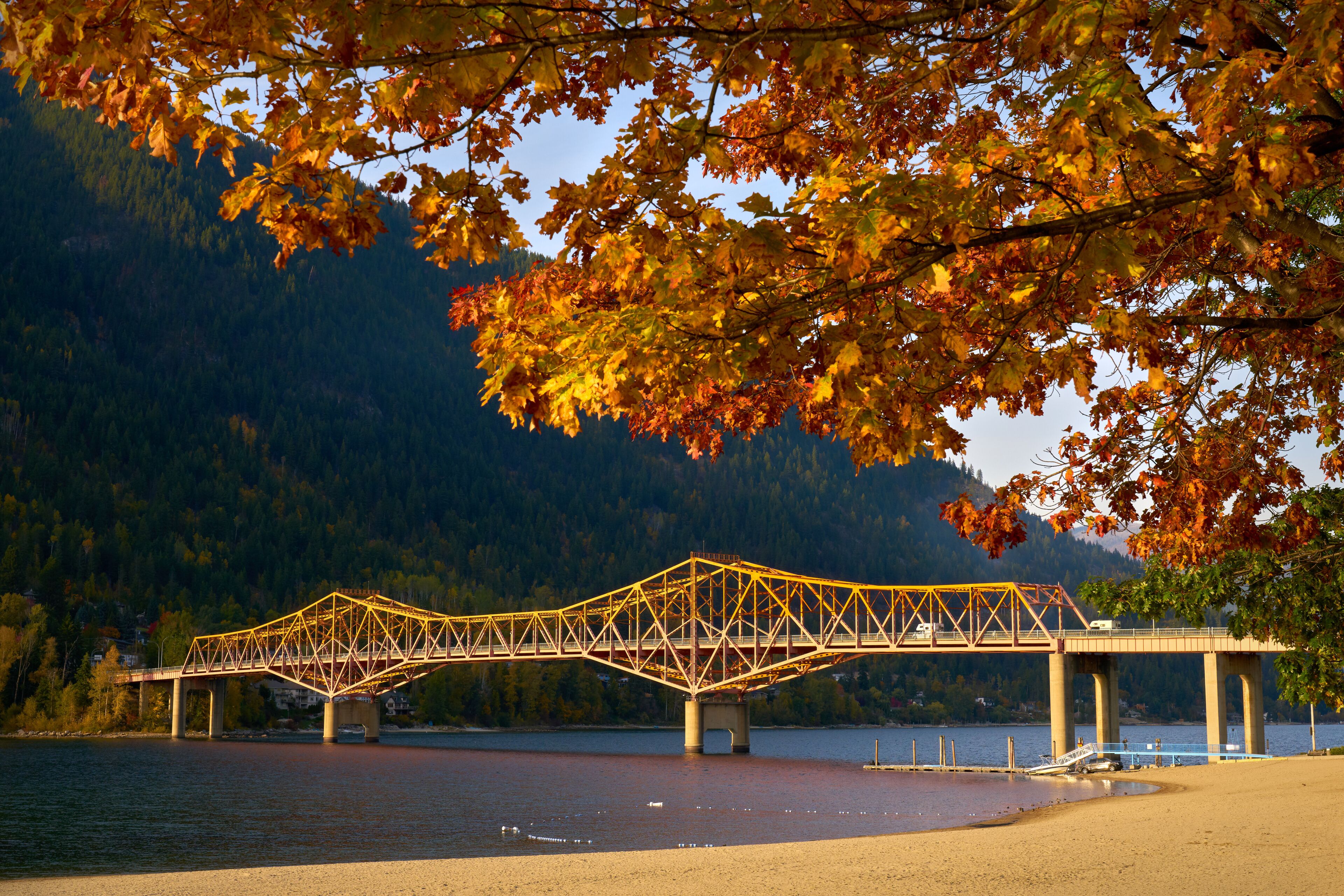 A steel truss bridge over a calm lake with autumn foliage in the foreground.