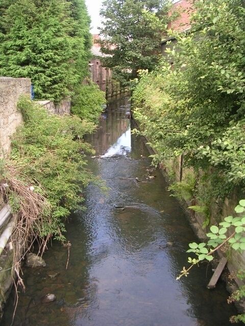 Um estreito canal de água com uma pequena cascata, rodeado por vegetação densa e muros de pedra.