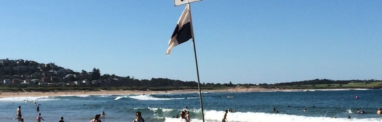Uma praia com um posto de salva-vidas, uma bandeira de salva-vidas, uma prancha de surf amarela, uma toalha de praia e uma cadeira de praia.