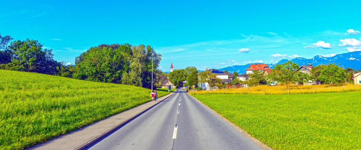 A rural road flanked by green fields, a church, and houses with mountains in the background.