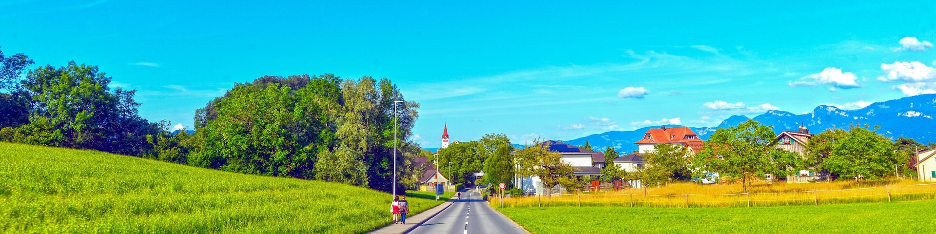 A rural road flanked by green fields, a church, and houses with mountains in the background.