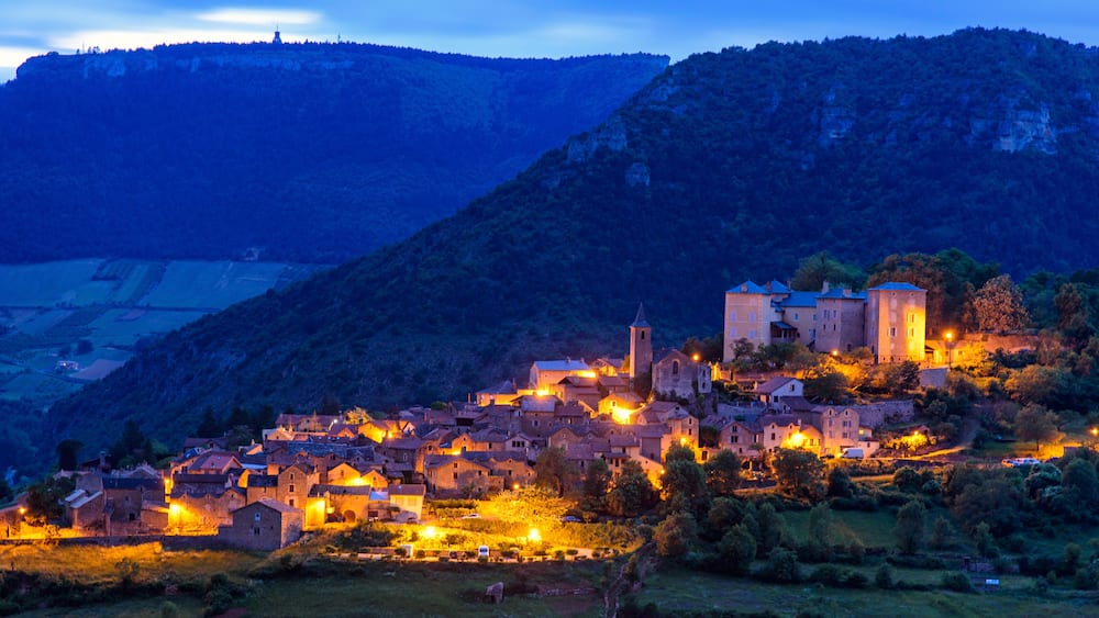 Un village au crépuscule, avec des bâtiments illuminés et une église imposante qui se dresse sur une colline.