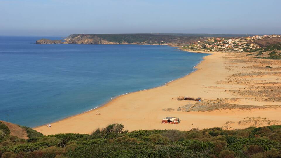 A coastal landscape with a sandy beach, a clear blue sea, and a distant town.