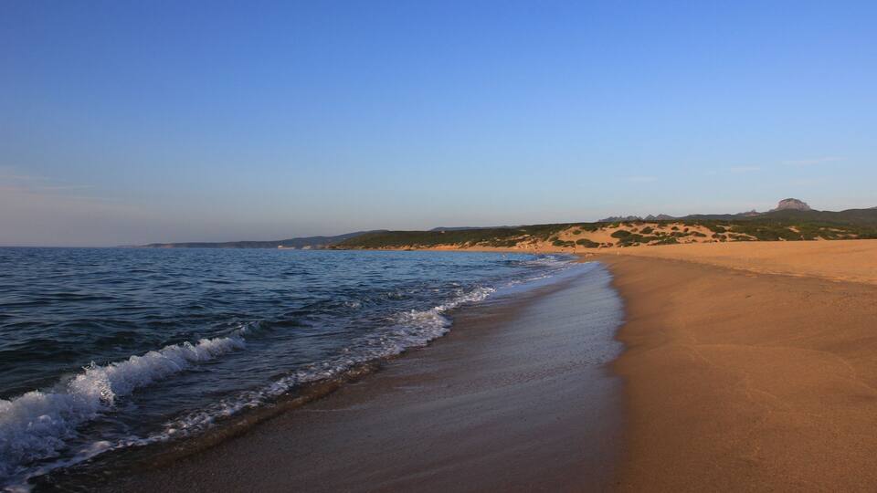 A beach with a clear sky, gentle waves, and a distant hill.