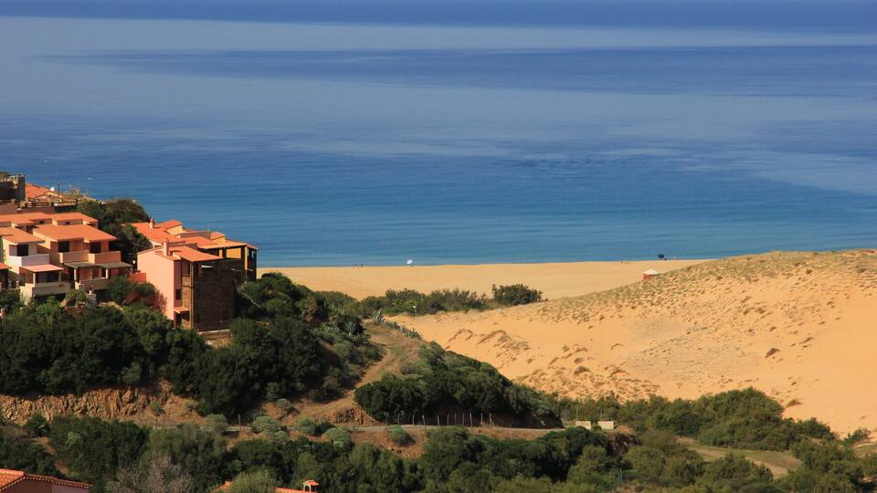 A coastal landscape with a sandy beach, a clear blue sea, and buildings with red-tiled roofs.