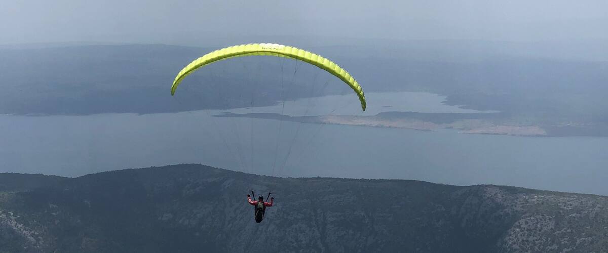 A paraglider with a yellow canopy flying over a mountainous landscape with a body of water below.