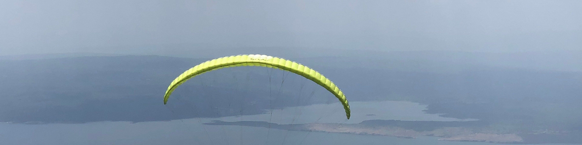 A paraglider with a yellow canopy flying over a mountainous landscape with a body of water below.