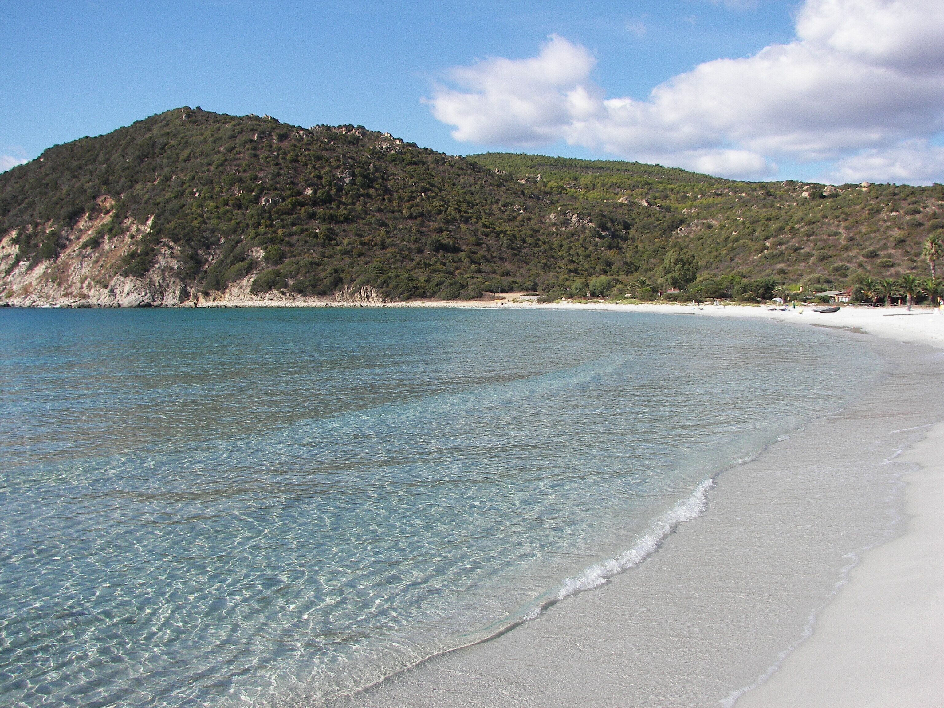 A clear beach with white sand and turquoise water, surrounded by a hilly landscape with sparse vegetation.