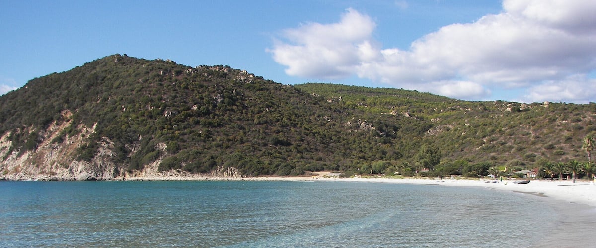 A clear beach with white sand and turquoise water, surrounded by a hilly landscape with sparse vegetation.
