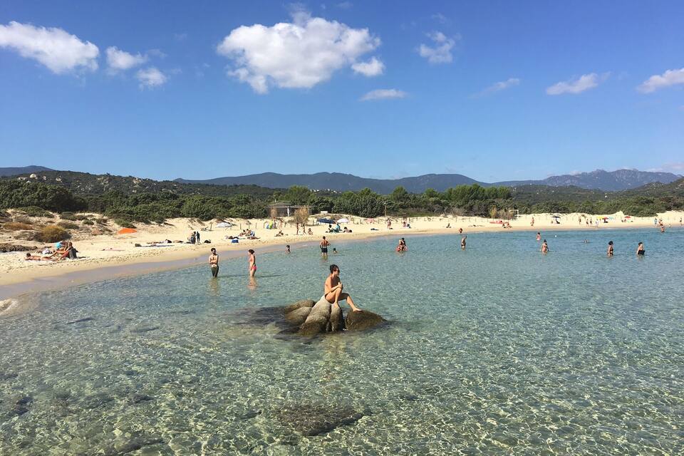 A beach with clear shallow waters, people swimming, and a woman sitting on a rock.