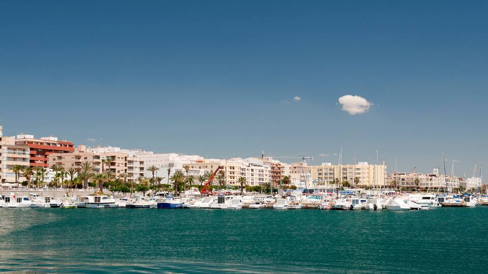 A marina with numerous boats docked, a clear blue sky, and a cityscape with buildings in the background.