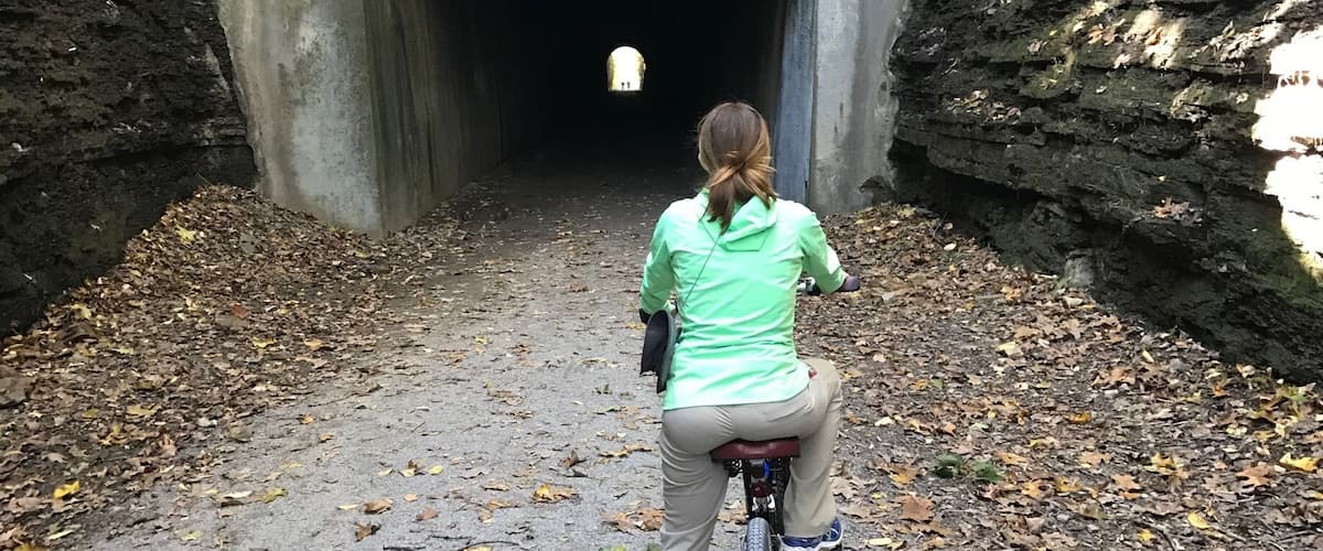 A person riding a bicycle towards a tunnel entrance surrounded by trees and fallen leaves.