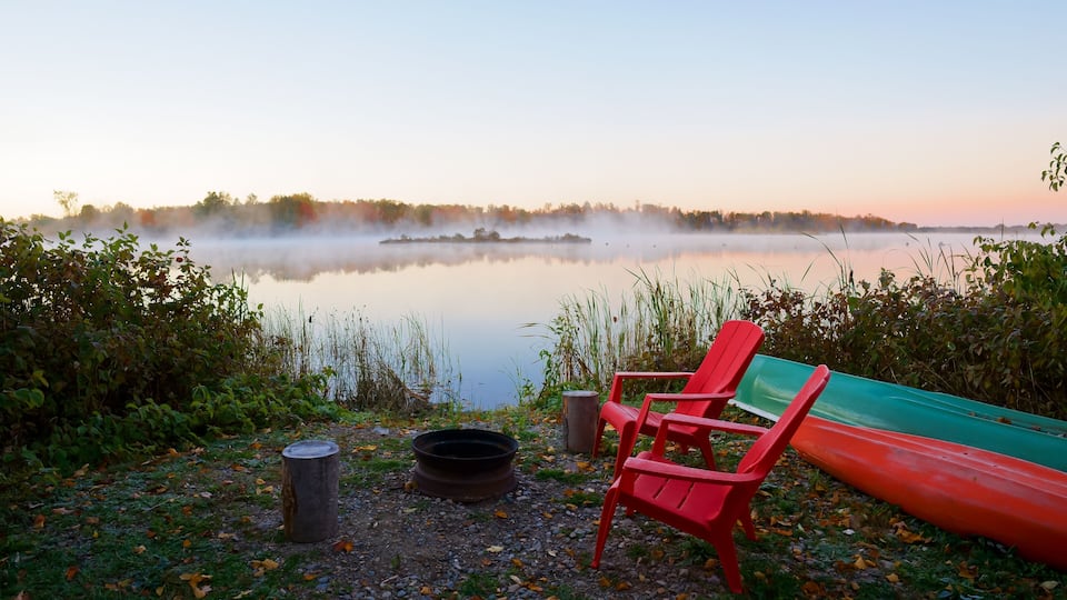 A serene lakeside scene with a red Adirondack chair, a green canoe, and a campfire pit.