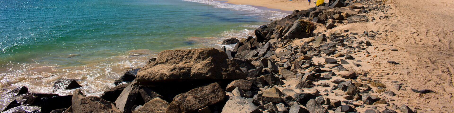 A rocky beach with clear blue water, a distant bridge, and a few people walking.