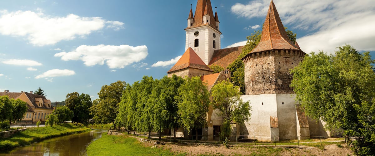 Eine historische Kirche mit einem hohen Turm und einem kleineren, daran grenzenden Wehrturm, umgeben von üppiger Vegetation und einem Fluss.