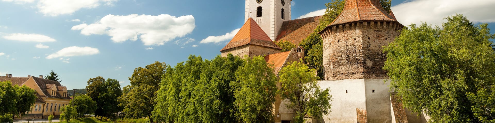 A historic church with a tall spire and a smaller adjacent tower, surrounded by lush greenery and a river.