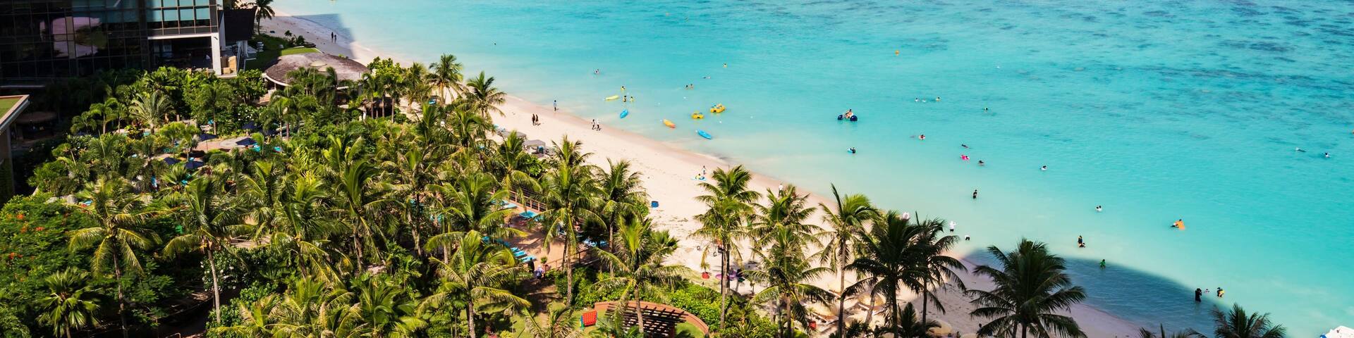 A tropical beach with clear turquoise waters, palm trees, and a resort area with swimming pools.
