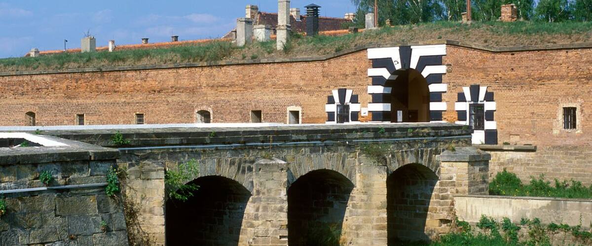 A stone bridge with arches over a river, surrounded by grassy fields and a brick wall with chimneys in the background.