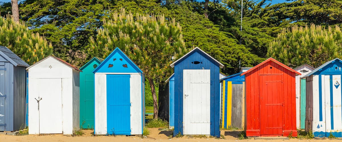 Colorful beach huts with blue, white, and red exteriors, set against a backdrop of lush green trees.