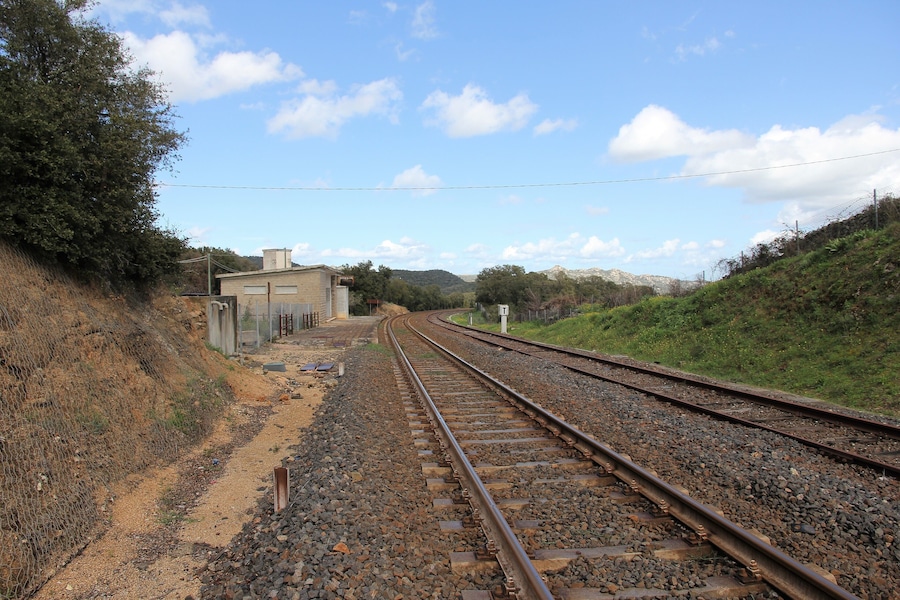 Vías de tren serpenteando por una zona montañosa con un edificio pequeño y una valla a la izquierda.