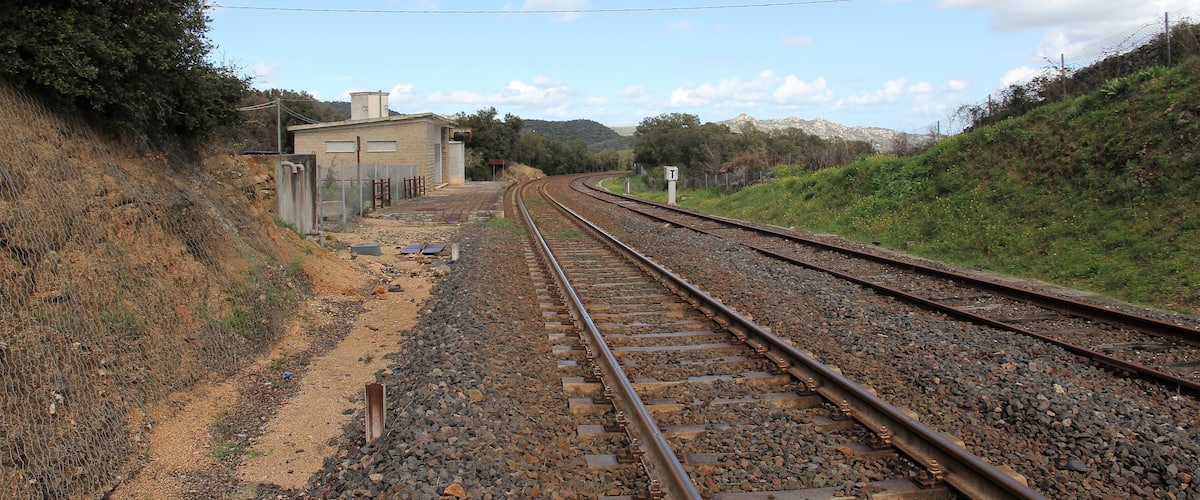 Railway tracks curving through a hilly area with a small building and a fence on the left.