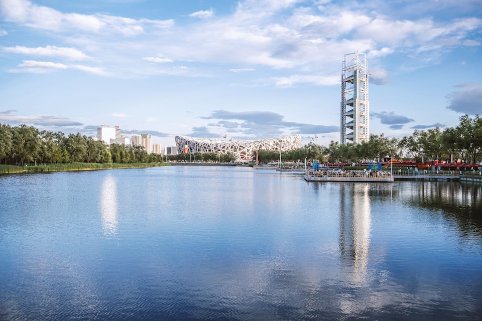A calm lake with a modern stadium and a tall tower in the background.